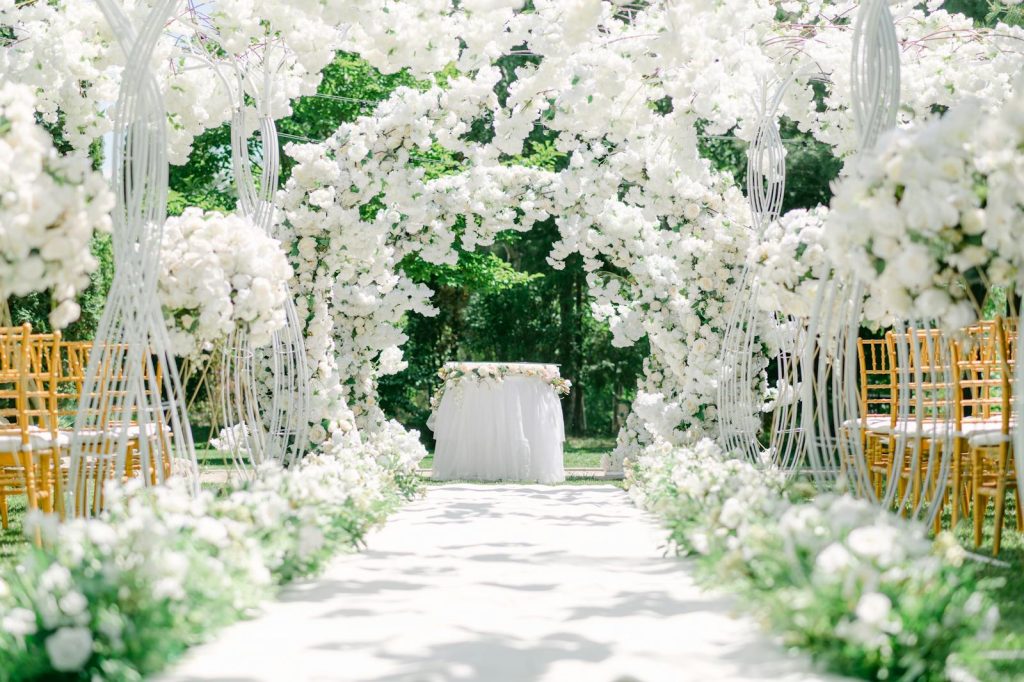 An elegant outdoor wedding venue setup with white floral arches and chairs.