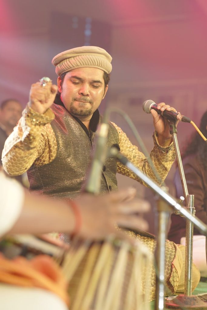 Traditional Indian singer performing live with microphone and tabla in vibrant setting.